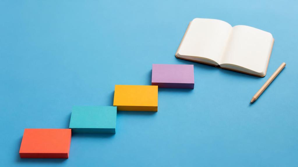 Four stacks of coloured sticky notes arranged like steps beside an open notebook and a pencil on a blue background