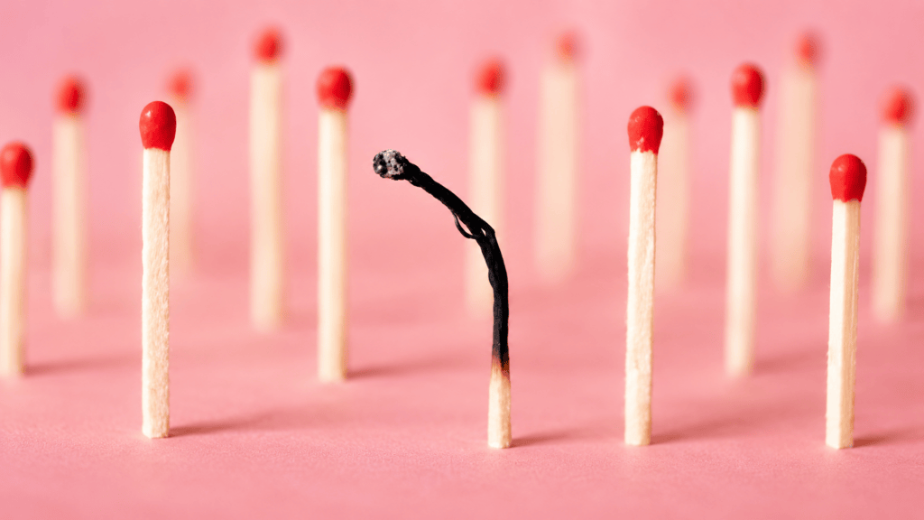 One burnt match among several unburnt red-headed matches on a soft pink surface.
