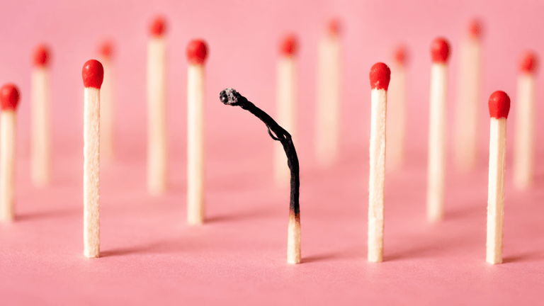 One burnt match among several unburnt red-headed matches on a soft pink surface.